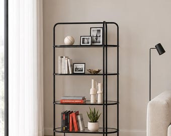 May include: A tall, four-tier shelving unit featuring a black metal frame with rounded corners and clear glass shelves. The shelves are filled with various decorative items, including books, framed photographs, a small potted plant, and vases.