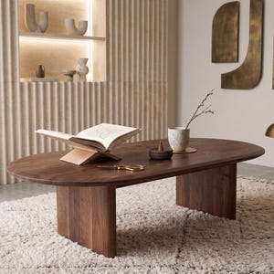May include: An oval dark wood coffee table with a smooth surface and two curved pedestal bases. On the table sits an open book on a wooden stand, a small dark bowl with an incense cone, a ceramic cup with a twig, and a pair of gold scissors.