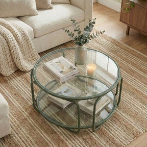 Puede incluir: Una mesa de centro redonda con estructura de metal verde salvia y dos niveles circulares de cristal. El nivel superior sostiene un jarrón de cerámica con ramas de eucalipto y una vela, mientras que el inferior muestra dos libros y un cuenco.