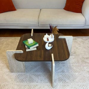 Puede incluir: Una mesa de centro cuadrada con una superficie de madera veteada de color marrón oscuro y cuatro patas de piedra de color beige claro. La mesa sostiene dos libros, una vela de burbujas blanca, una bandeja circular blanca con recipientes y flores.