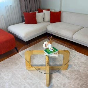 May include: A coffee table with a clear glass top and a light-toned wooden base featuring a cross-shaped design. The table holds a small white dish with decorative items and a green book. The table is positioned in front of a large sectional sofa.