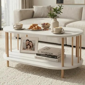 May include: An oval white coffee table with two tiers and light wood cylindrical legs. The top surface holds two ceramic mugs, a plate of croissants, a bowl of nuts, and a vase with greenery. The bottom shelf displays a photo frame and books.