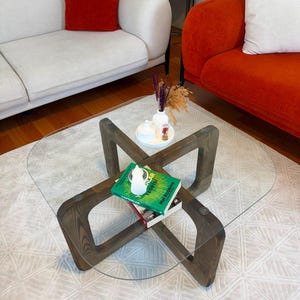 May include: A coffee table with a clear square glass top and a dark brown wooden base featuring an intersecting cross design. On the table sits a white circular tray with a small white vase containing dried flowers, and two stacked books.