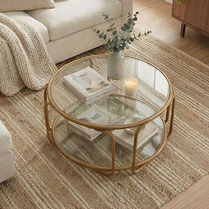 May include: A round coffee table with a gold-toned metal frame and two clear glass tiers. The top tier holds a light-colored ceramic vase with green eucalyptus stems and a small white candle, while the bottom tier displays two stacked books.
