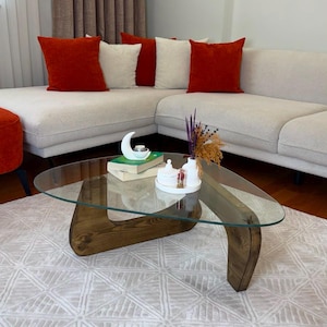 May include: A coffee table with a triangular glass top and a dark brown wooden base featuring curved legs. The table holds two stacked books, a white crescent moon-shaped object, a small white lidded container, and a white vase with dried flowers.