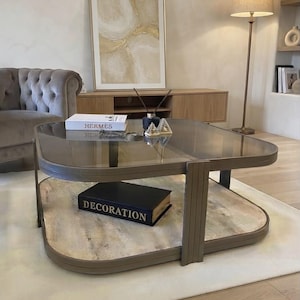 May include: A square coffee table with a dark-tinted glass top and a lower shelf featuring a light-coloured stone surface. The frame is constructed from a metallic material with a muted bronze finish and rounded corners, supporting the two-tiered design.