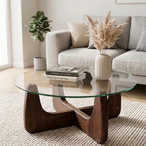 May include: A round coffee table with a clear glass top and a dark brown wooden base featuring intersecting curved legs. The table holds a white ribbed vase with dried pampas grass, a small gray geometric object, and two stacked books.