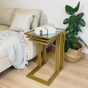 Könnte beinhalten: Ein Set aus drei Satztischen mit goldfarbenen Metallrahmen und rechteckigen Glasplatten. Die Tische sind ineinander gestapelt und zeigen ein gestuftes Design mit klaren Linien und einer metallischen Oberfläche für Ablagefläche.