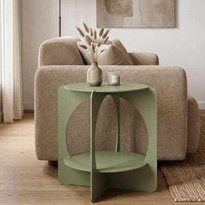 Puede incluir: Una mesa auxiliar de color verde salvia con una superficie circular y un estante inferior sostenido por cuatro paneles verticales con recortes arqueados. La mesa está construida con piezas planas entrelazadas, creando una estructura geométrica.