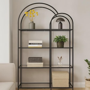 Puede incluir: Una estantería de metal negro con estantes de cristal y dos secciones superiores arqueadas. Los estantes contienen un jarrón gris con flores amarillas, un libro titulado Sapiens, una lámpara oscura, una planta verde y dos cajas de almacenaje.