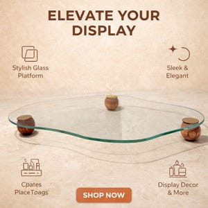 Puede incluir: Una plataforma de exhibición de vidrio transparente con forma orgánica, apoyada sobre tres pies de madera redondos con tapas doradas. La imagen incluye el texto: ELEVATE YOUR DISPLAY, Stylish Glass Platform, Sleek & Elegant, Creates Place Trays, Display Decor & More, SHOP NOW.
