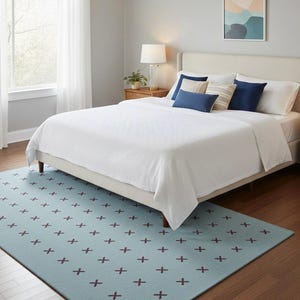 Puede incluir: Una alfombra rectangular con un color base azul claro y un patrón repetitivo de pequeños símbolos de cruz de color marrón oscuro dispuestos en cuadrícula. La alfombra se sitúa sobre un suelo de madera bajo una cama con ropa blanca y cojines azules.