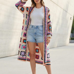 Könnte beinhalten: Eine lange, offene Strickjacke mit einem bunten Granny-Square-Häkelmuster. Das Kleidungsstück besteht aus cremefarbenem Garn mit Akzenten in Blau, Pink, Orange, Lila und Schwarz, ausgestattet mit langen Ärmeln und einem geraden Saum.