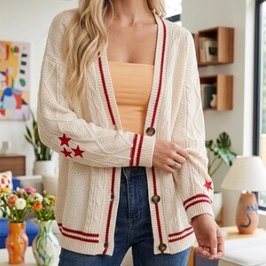 May include: A cream-colored cable knit cardigan featuring a V-neckline, long sleeves with red star motifs, and red striped trim along the button placket and cuffs. The garment is secured with four brown buttons down the front center.