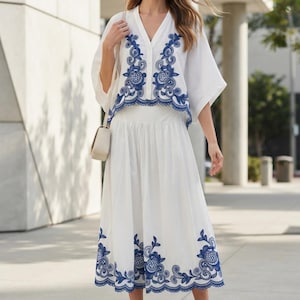 May include: A white two-piece set consisting of a V-neck blouse with short wide sleeves and a midi skirt. Both pieces feature intricate blue floral embroidery along the edges. The blouse has a button-down front and the skirt has a flared silhouette.