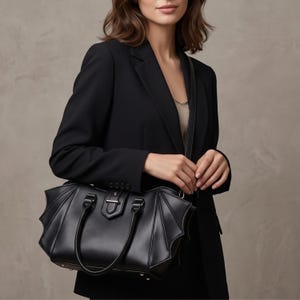 Può includere: Una borsa a mano nera dal design strutturato con pannelli laterali a forma di ali di pipistrello. La borsa include due manici superiori, una tracolla rimovibile e una chiusura centrale in metallo color argento sulla patta frontale.