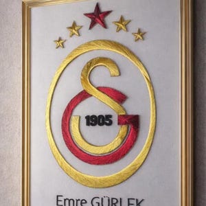 Puede incluir: Una pieza de arte en hilo enmarcada que presenta el logotipo del club deportivo Galatasaray en hilo amarillo y rojo. El diseño incluye un borde ovalado, las letras G y S, el año 1905 en negro, cuatro estrellas arriba y el nombre Emre GÜRLEK en negro.