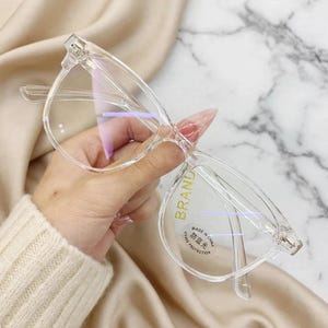 Puede incluir: Gafas con montura transparente y lentes con filtro de luz azul. El cristal derecho tiene una pegatina dorada que dice BRAND y una pegatina circular con el texto MADE IN CHINA, UV400 PROTECTION y caracteres chinos de luz azul.
