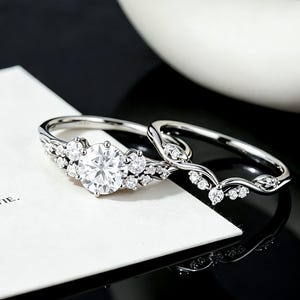 May include: A silver-toned engagement ring set featuring a large round center stone flanked by smaller accent stones, paired with a curved wedding band adorned with a series of tiny round stones along its delicate, flowing metal band.