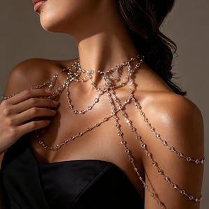 May include: A silver-toned body chain necklace featuring multiple delicate strands adorned with small, clear, round-cut crystals. The jewellery drapes across the neck, chest, and shoulders, creating a layered, intricate design with metallic links.