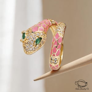Peut inclure: Une bague serpent dorée ornée d'écailles en émail rose, de cristaux clairs sertis pavé sur la tête et le corps, et de deux pierres vertes taille marquise pour les yeux. La bague a un design ouvert et inclut le texte Solac Jewelry Co en bas.