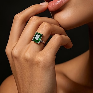 Pode incluir: Um anel de tom prateado com uma grande pedra preciosa verde de corte esmeralda retangular, fixada por quatro garras. A banda é lisa e polida, apresentando um design limpo que destaca a cor verde profunda e vibrante da pedra central.