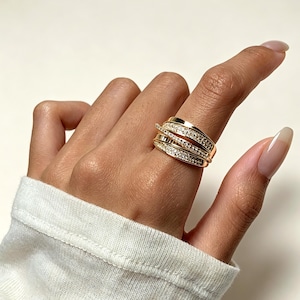 May include: A gold-toned ring featuring a multi-band crossover design. The ring consists of three distinct bands that intersect, with two of the bands encrusted with small, clear, sparkling stones along their entire visible surface.