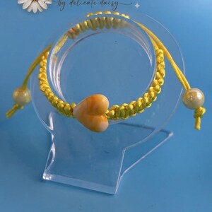 Può includere: Un braccialetto in cordino giallo con una perla centrale a forma di cuore con motivi marmorizzati arancioni e bianchi. Il braccialetto include due perle rotonde più piccole alle estremità dei cordini regolabili. Il testo "Beads by delicate daisy" è visibile.
