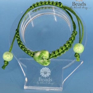 Può includere: Un bracciale in cordino intrecciato verde con una perla centrale a forma di cuore con motivi marmorizzati bianchi e verdi. Due perle sferiche più piccole di colore verde chiaro sono fissate alle estremità del cordino regolabile. Il testo Beads by delicate daisy è visibile.
