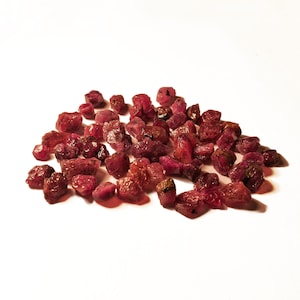 May include: A collection of small, raw, unpolished ruby gemstones in various shades of deep red and magenta. Each stone features an irregular, jagged shape with natural facets and textures, clustered together to form a dense, scattered pile.