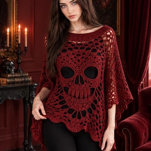 May include: A dark red crochet poncho featuring an intricate skull pattern throughout the knit design. The garment has a wide, rounded neckline and a loose, flowing silhouette with scalloped edges, worn over a solid black base layer.