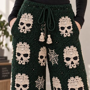 May include: Dark green crochet pants featuring a repeating pattern of cream-colored skull and spiderweb motifs. The pants have a wide-leg silhouette, an elasticized waistband with a matching dark green drawstring, and small cream-colored tassels.