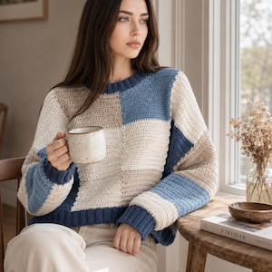 May include: A long-sleeve sweater featuring a color-blocked pattern of cream, beige, and blue squares. The garment has a ribbed crew neckline, ribbed cuffs, and a ribbed hem, constructed with a visible crochet stitch texture throughout the body.