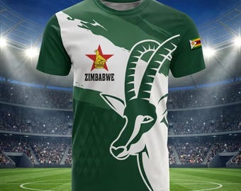 May include: A short-sleeved t-shirt featuring a green and white colour scheme with a large graphic of an antelope head. The design includes a red star with a yellow bird emblem, the text ZIMBABWE, and a small Zimbabwe flag on the sleeve.