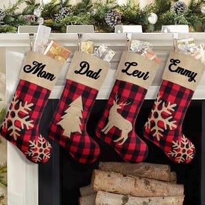 May include: Four Christmas stockings with red and black buffalo plaid fabric and tan burlap cuffs. The cuffs are personalized with the names Mom, Dad, Levi, and Emmy in black script. Each stocking features a tan burlap applique of a snowflake or tree.