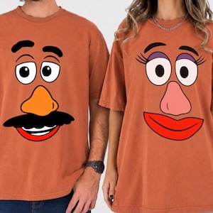 May include: Two rust-colored crew neck t-shirts featuring cartoon face graphics. The left shirt displays a face with black eyebrows, large white eyes, an orange nose, and a black mustache. The right shirt shows a face with long eyelashes, purple eyeshadow, a pink nose, and red lips.