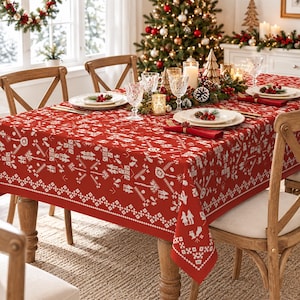 May include: A rectangular red tablecloth featuring a white Nordic-inspired pattern of geometric shapes, trees, and figures. The fabric covers a wooden dining table set with white plates, clear glasses, and red napkins, creating a festive display.