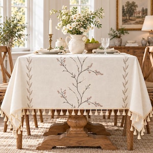 May include: A rectangular off-white tablecloth featuring a central embroidered pattern of thin brown branches with small pink blossoms. The edges are finished with a decorative trim of small, dangling cream-colored tassels on all sides.