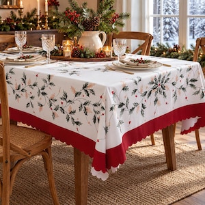 May include: A white rectangular tablecloth featuring a printed pattern of green and gold holly leaves with red berries. The tablecloth has a wide solid red border along the bottom edge with a scalloped hemline, draped over a wooden dining table.