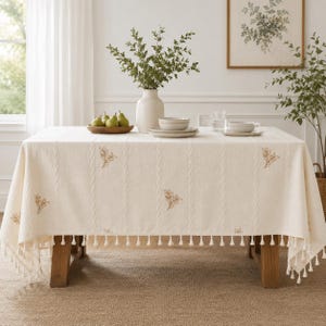 May include: A cream-colored rectangular tablecloth featuring vertical cable knit-style textures and small embroidered brown leaf motifs. The edges are finished with a decorative fringe of small white tassels hanging along the perimeter.