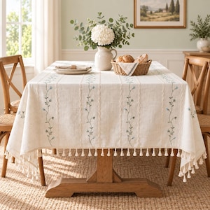 May include: A cream-coloured rectangular tablecloth featuring vertical embroidered green vines with small blue flowers and a braided texture pattern. The edges are finished with a decorative fringe of small white tassels hanging from the hem.