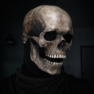 Op de afbeelding: Een realistisch menselijk schedelmasker met verweerde botkleurige texturen, diepe donkere oogkassen en een open kaak met zichtbare tanden. Het masker wordt gedragen over een effen zwarte stoffen capuchon die de nek en schouders bedekt.