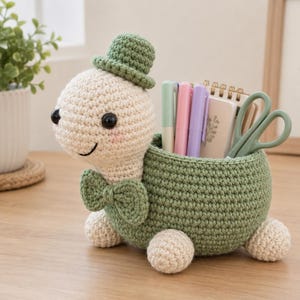 Può includere: Un organizer da scrivania all'uncinetto a forma di tartaruga con testa color crema, occhi neri, cilindro verde e papillon abbinato. Il guscio è una ciotola verde scuro che contiene diverse penne, un piccolo taccuino e un paio di forbici.
