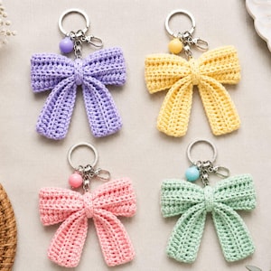 Può includere: Quattro portachiavi a forma di fiocco all'uncinetto nei colori lavanda, giallo, rosa e verde menta. Ogni fiocco presenta un anello in metallo argentato, un moschettone e una piccola perlina rotonda abbinata fissata al centro del fiocco.