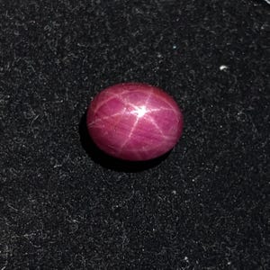 Könnte beinhalten: Ein tief magentafarbener, ovaler Sternrubin-Edelstein mit einem ausgeprägten sechsstrahligen Asterismus-Effekt, der von der Mitte der glatten, polierten Cabochon-Oberfläche ausgeht und ein helles weißes Sternmuster auf dem dunkelrosa Stein bildet.