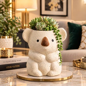 May include: A cream-colored, speckled ceramic planter shaped like a koala, featuring a dark brown nose and two small black eyes. The planter holds a cluster of green succulents and a trailing string of pearls plant that hangs down the right side.