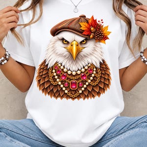 May include: A white t-shirt featuring a graphic of a bald eagle wearing a brown tweed flat cap adorned with autumn leaves and acorns. The eagle is also wearing a layered gold chain necklace with white pearls and large, faceted red gemstones.