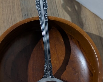 Könnte beinhalten: Eine silberfarbene Metallschöpfkelle mit einem verzierten, geprägten Blumenmuster am Griff. Der Griff endet in einem dekorativen, abgerundeten Oberteil mit komplizierten Schnörkeln und Blütenblättern, während die Laffe glatt und reflektierend ist.