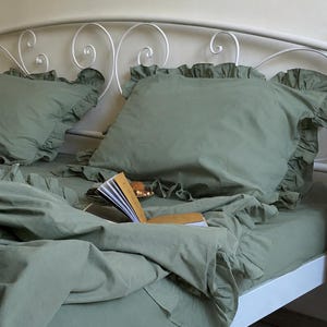 May include: A sage green bedding set featuring two ruffled pillowcases and a matching duvet cover. The bedding is displayed on a white metal bed frame. An open book with yellow pages rests on the duvet, partially obscuring a small plate of snacks.