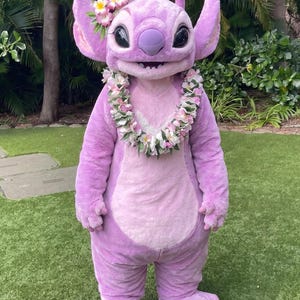 Peut inclure: Un costume en peluche intégrale représentant un personnage extraterrestre rose avec de grandes oreilles, un nez large et une bouche souriante. Le personnage porte un collier de fleurs autour du cou et des fleurs blanches et roses derrière l'oreille.